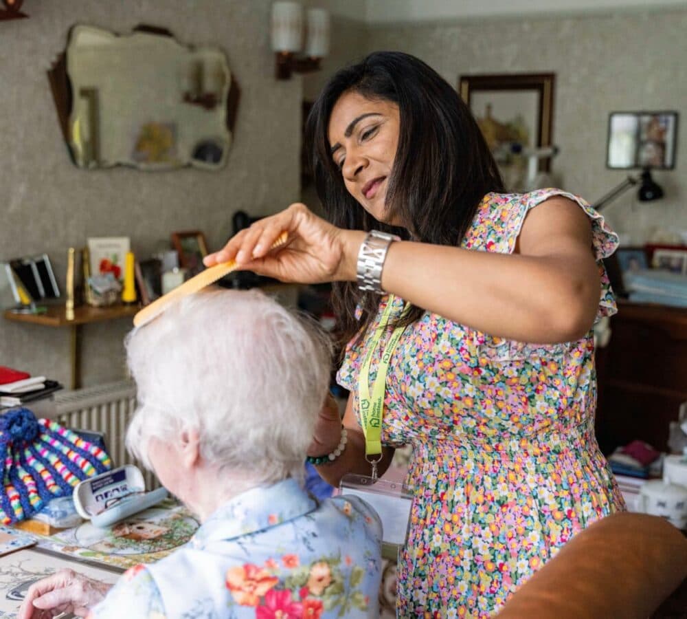 A female carer with long black hair combing the hair of an older female adult