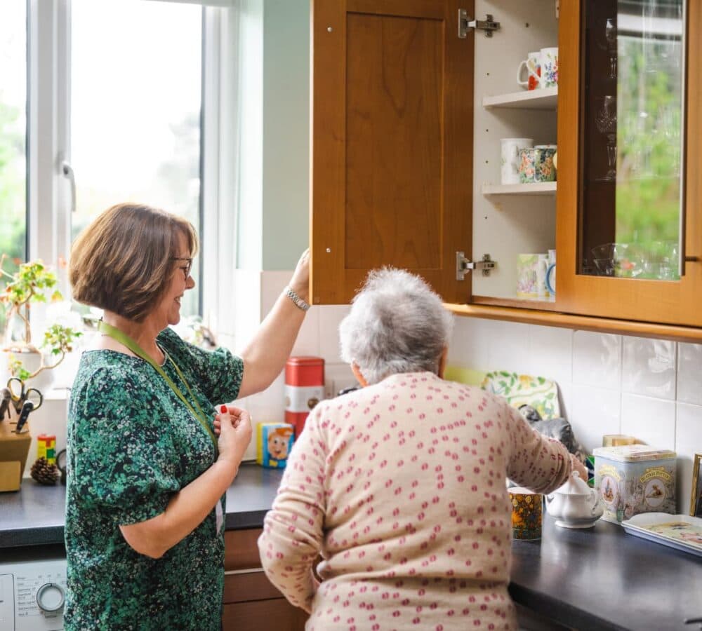 An older female adult with grey hair doing something in the kitchen with the help of a younger female with short hair and wearing green