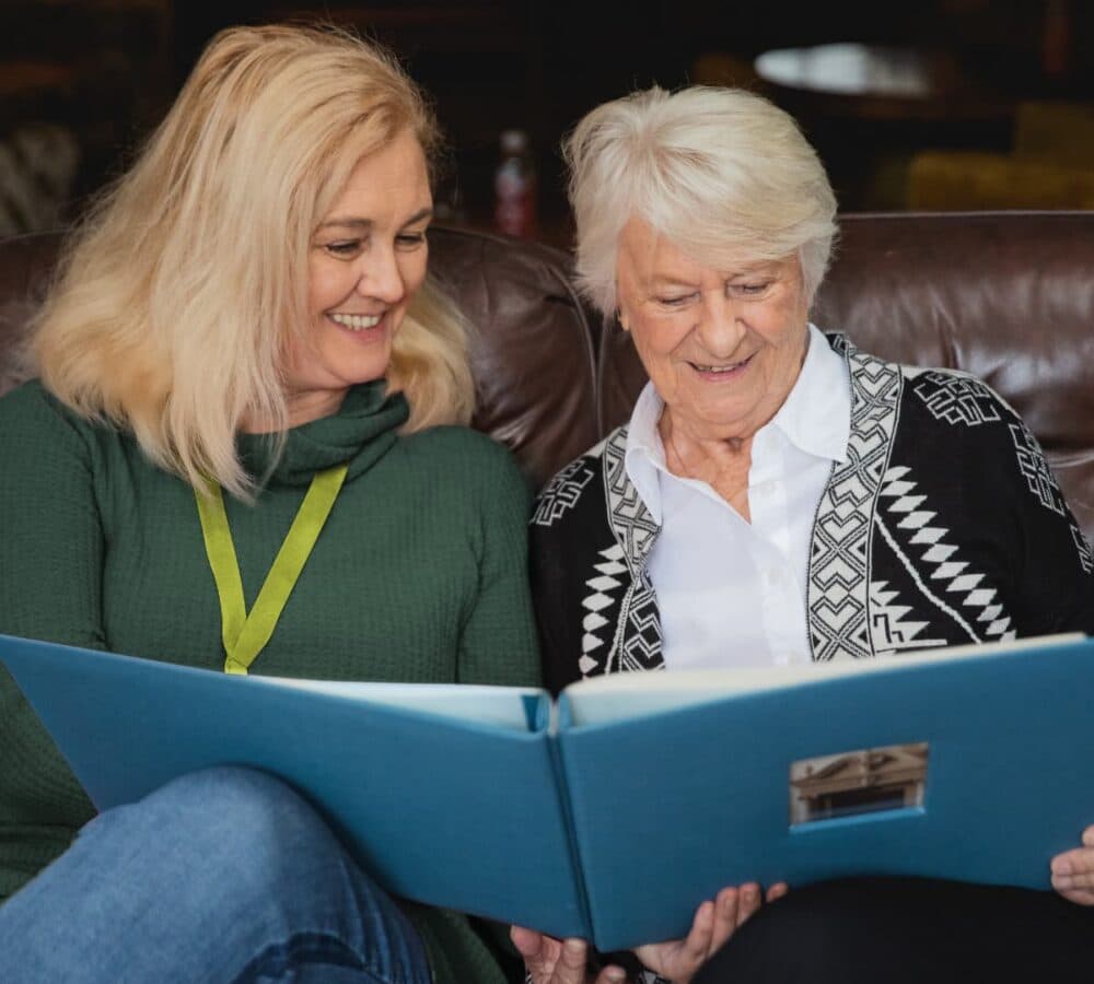 An older female adult looking at a photo album with her younger female carer with blonde hair both sitting and smiling inside the house - Home Instead