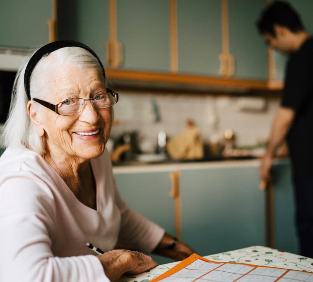 An older female adult with white hair and wearing eyeglasses happy and smiling while sitting in the kitchen