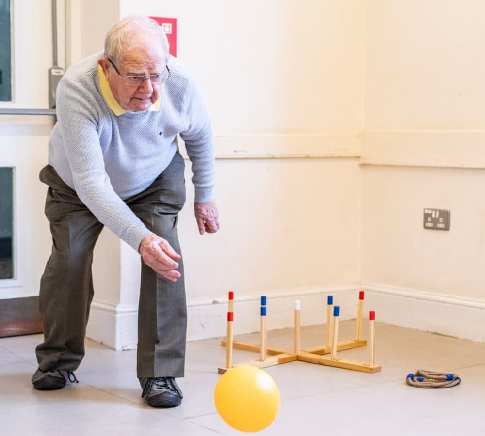 An older male adult with grey hair and wearing eyeglasses, playing ball inside the room