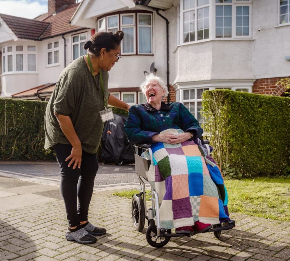 An older female adult with short white hair sitting on a wheelchair with her younger female carer with short black hair and wearing green both happy and smiling while walking outdoors