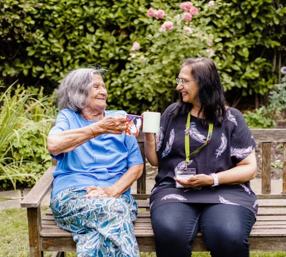 Two women having coffee while sitting on the bench in the garden both happy and smiling