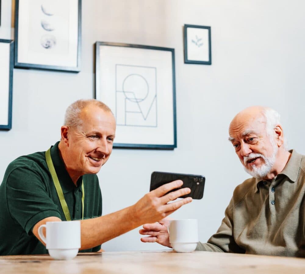 An older male adult with white hair watching on the phone with his younger male carer while drinking coffee
