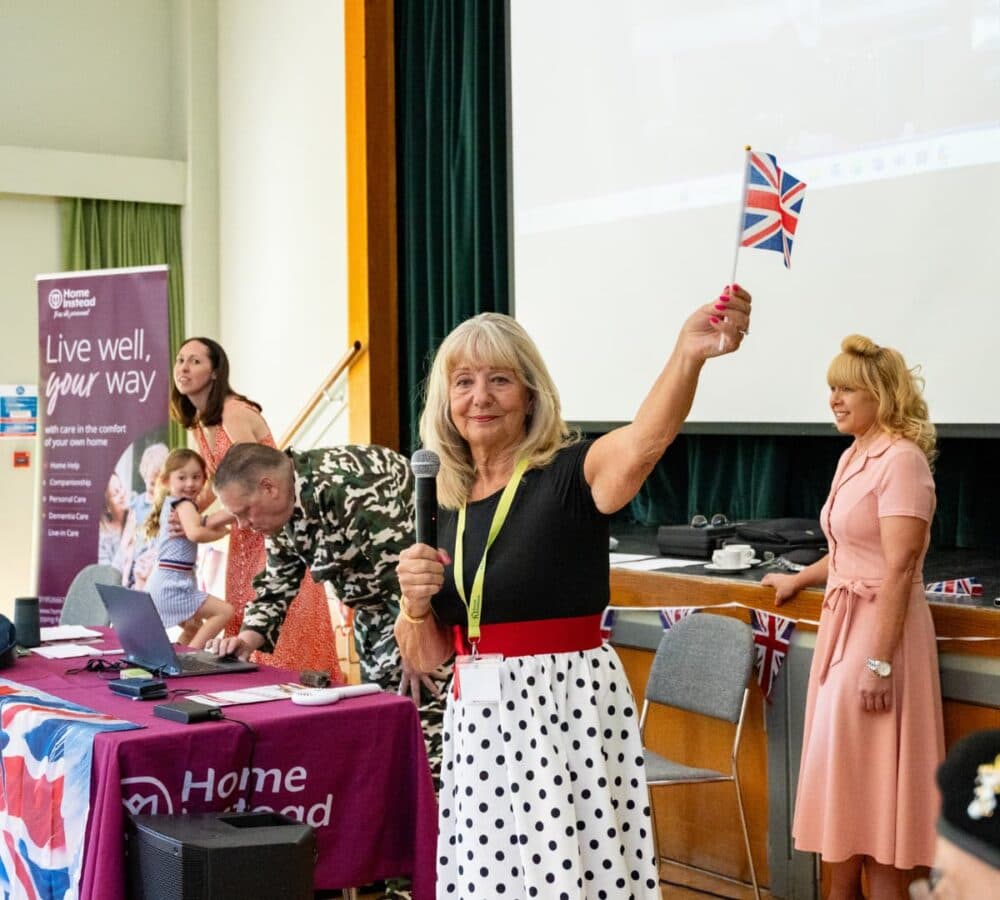An older female adult with long blonde hait standing and smiling while wearing black top and polka dot skirt and waving a flag inside the hall