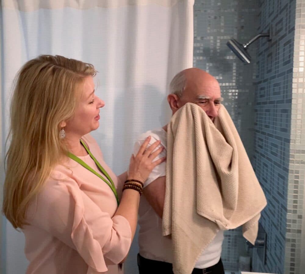 An older male adult with grey hair wiping his face using a towel with the hep of his younger female carer with long blonde hair and wearing pink inside the bathroom
