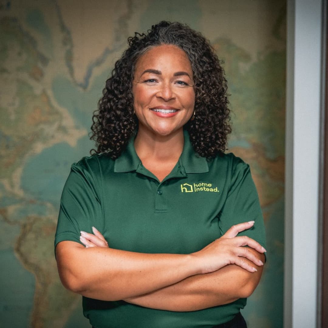 Woman in a green Home Instead shirt stands smiling with arms folded in front of a world map background. - Home Instead