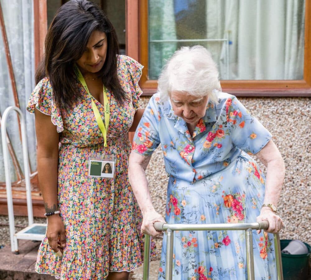 An older female adult with white hair going outside while using a walker and being help by her younger female carer with long black hair