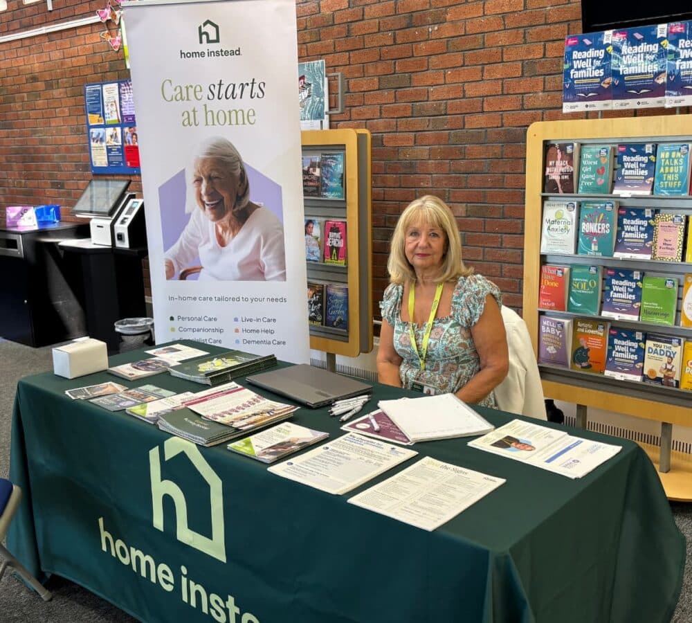 A woman with long blonde hair and wearing green sitting while having some flyers on the table happy and smiling