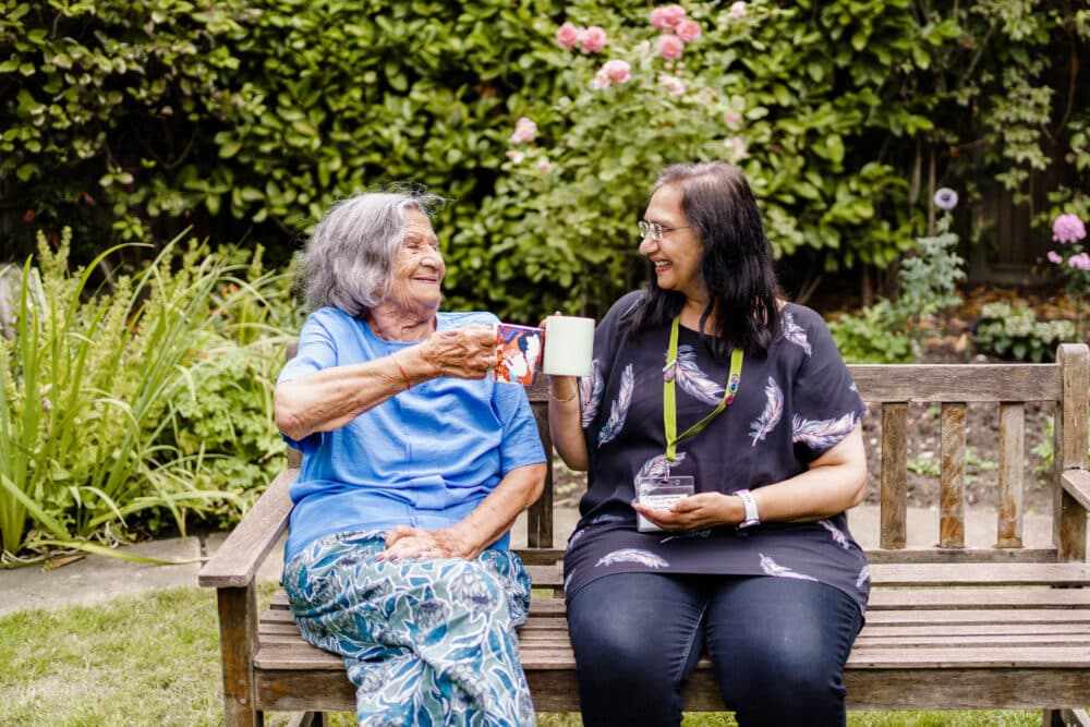 Two women having coffee together while sitting on a bench both happy and smiling in the garden
