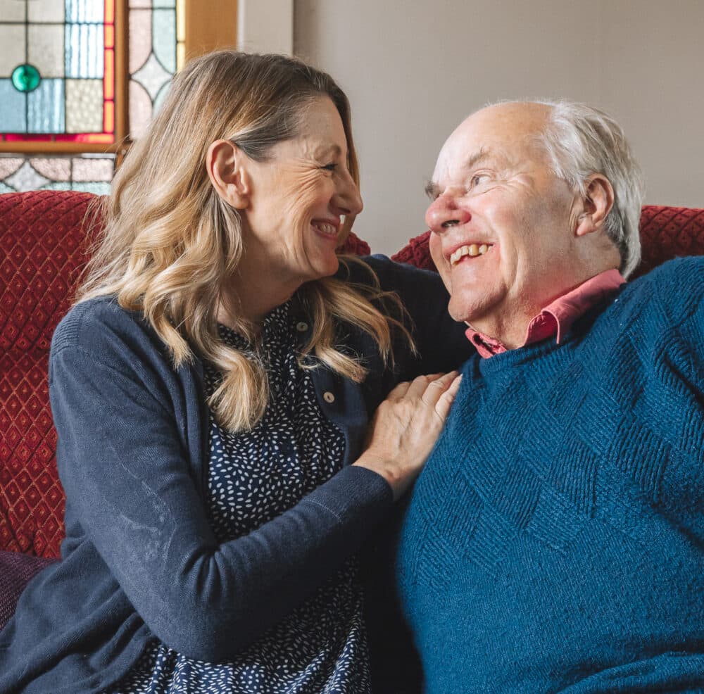 A woman sits next to an older man on a sofa. They smile at each other and she holds his shoulder.
