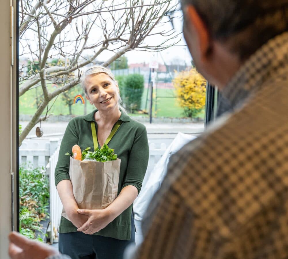 A woman holding a bag of groceries greeting an older male adult at the door of the house