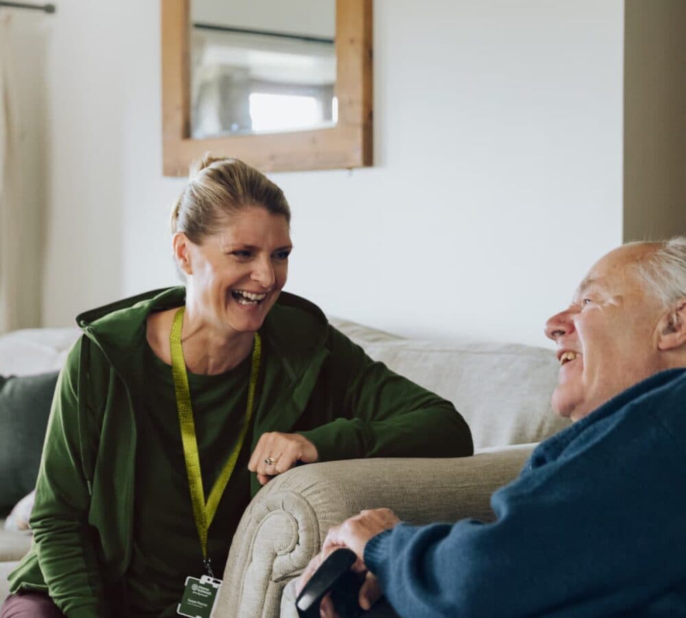 An older male adult chatting with his carer while sitting on the couch both happy and smiling