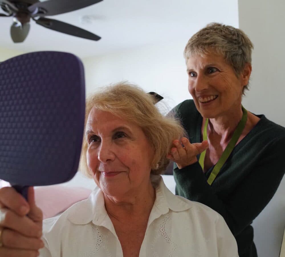 An older woman with short grey hair looking at the mirror while being combed by her younger female carer with short hair and wearing blue
