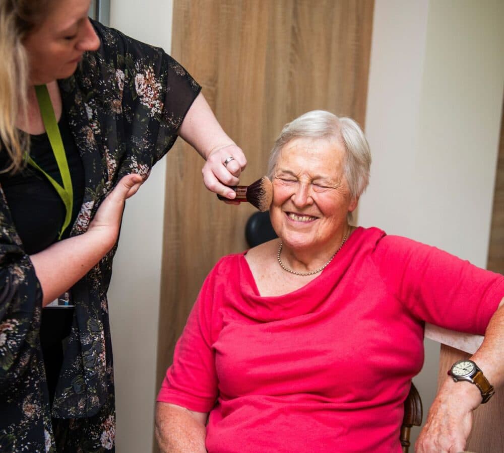 An older female adult with short hair happy and smiling while sitting on a chair and having her make up on by her younger female carer