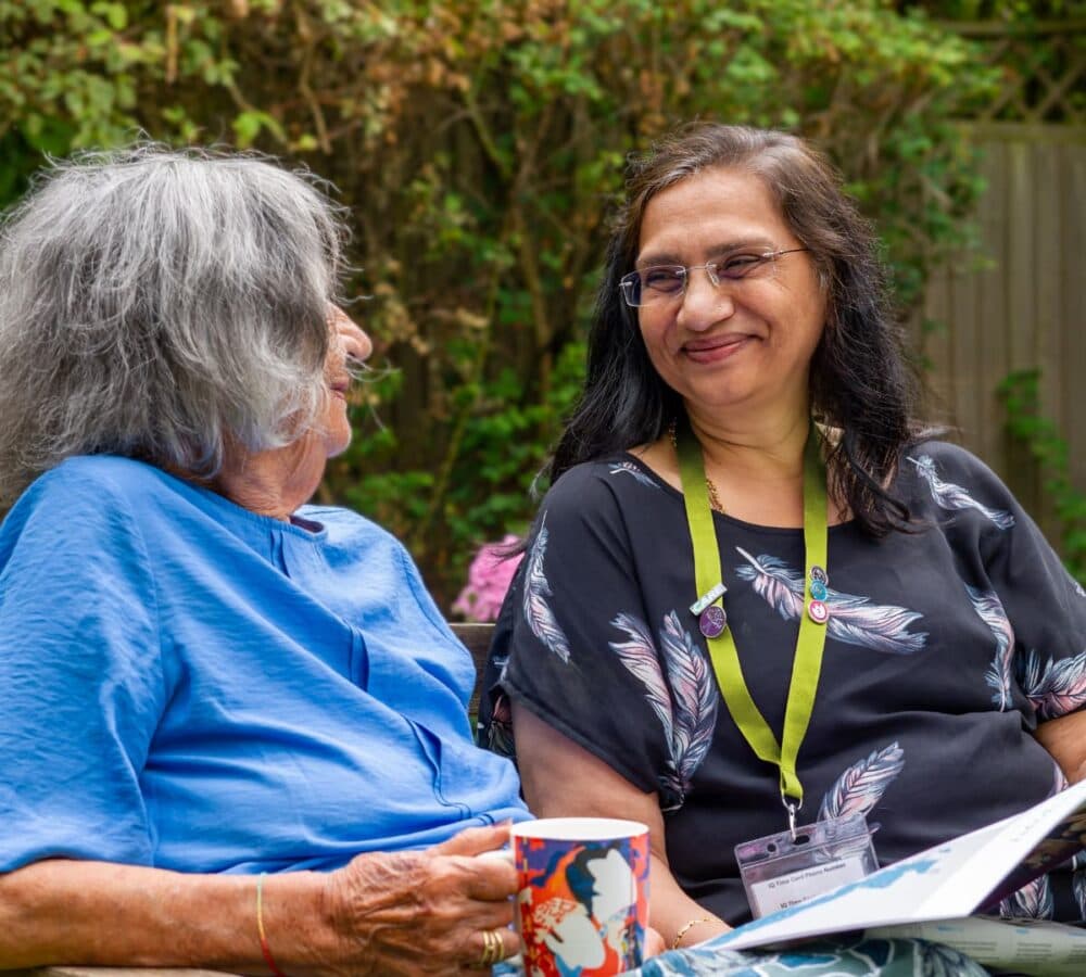 Two women chattung and drinking coffee while sitting on a bench in the garden