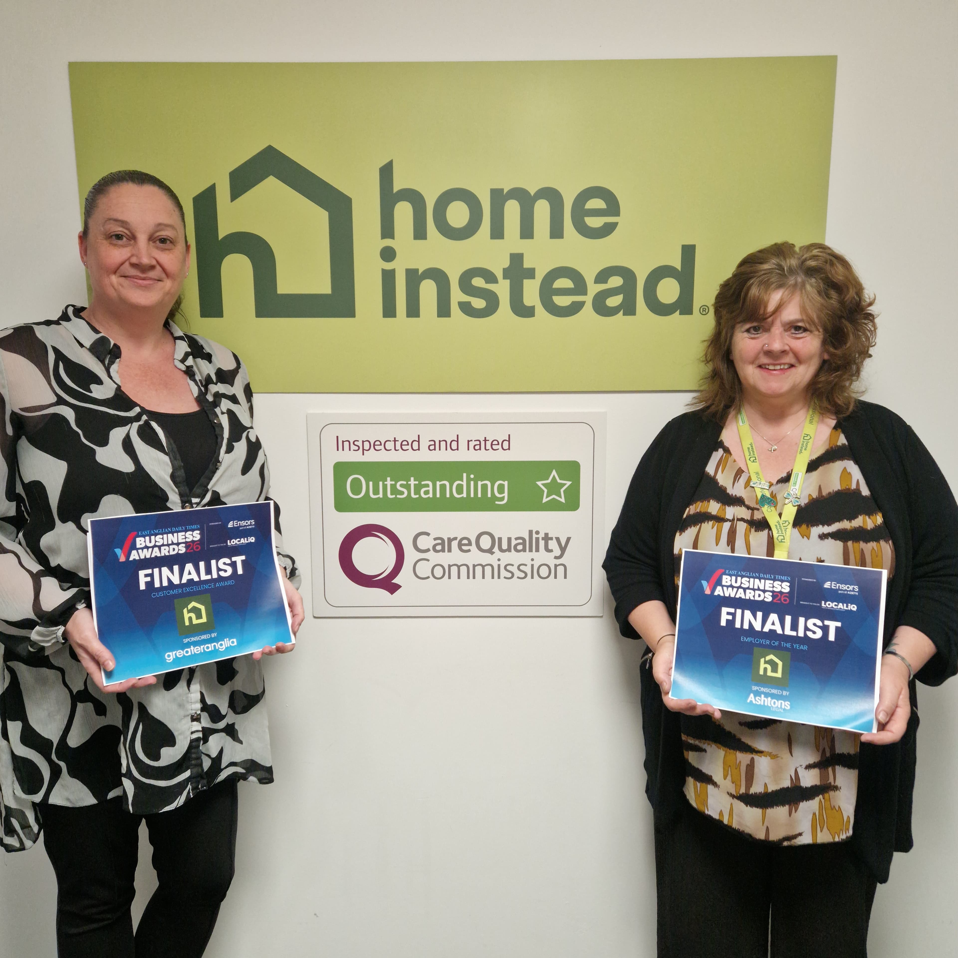 Two women holding "Business Awards Finalist" signs stand in front of a Home Instead logo and award sign. - Home Instead