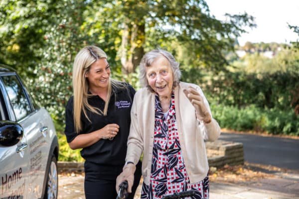 Smiling carer helps an elderly woman with a walking frame outdoors beside a car on a sunny day. - Home Instead