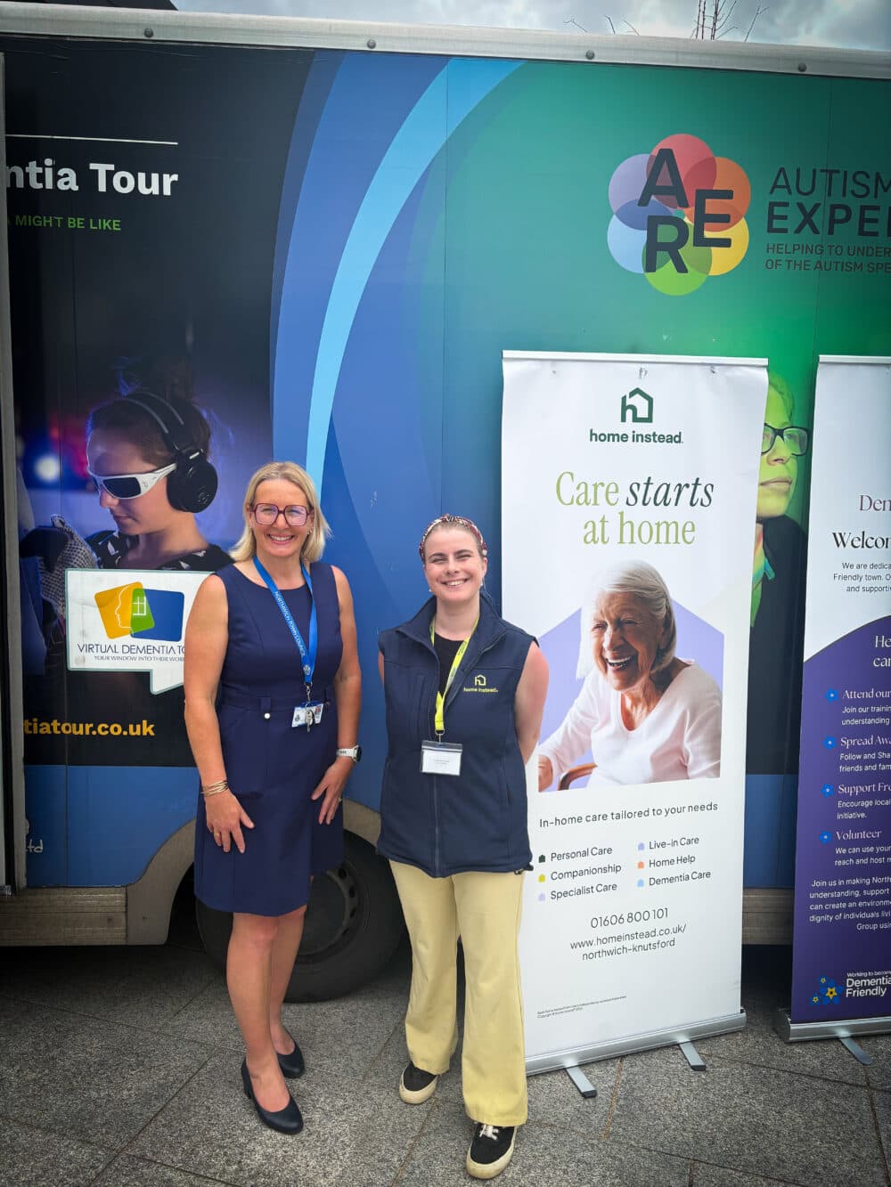 Two women stand smiling in front of a care home banner and an Autism Experience tour van. - Home Instead