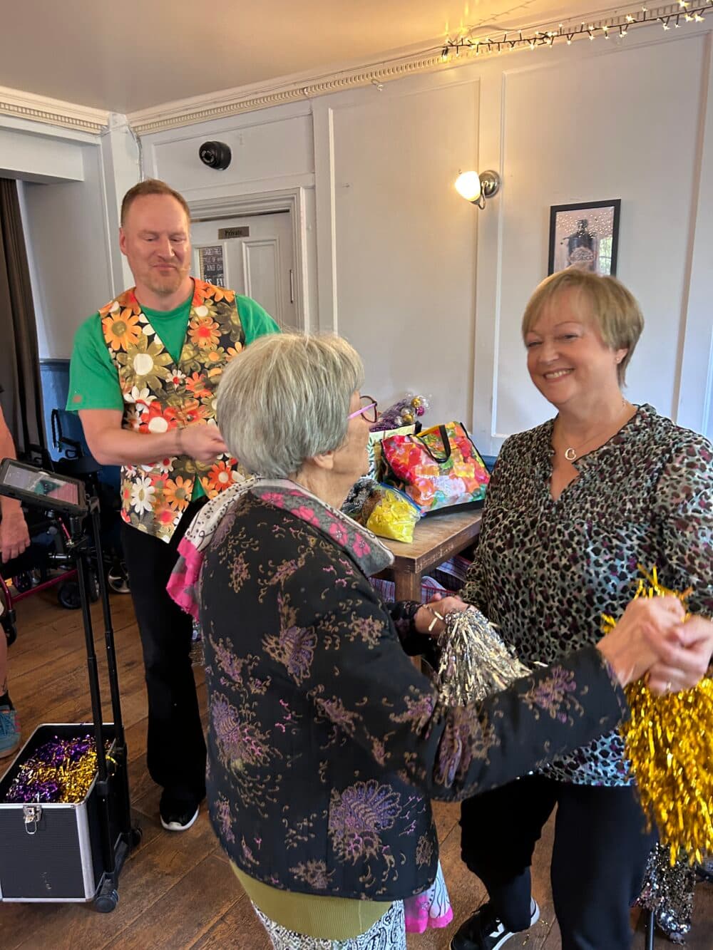 An older woman and a smiling woman dance together with colourful pom-poms during a lively community singing session, while an entertainer performs in the background.