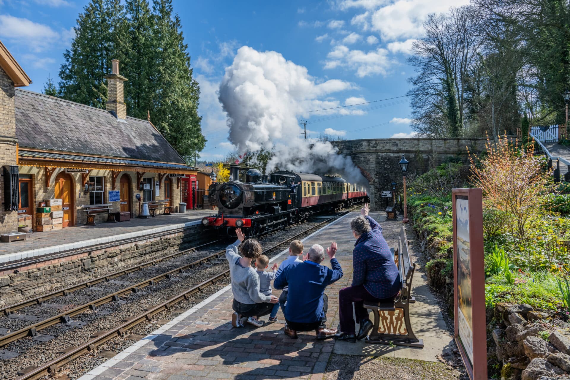 A steam train arrives at a quaint station as four people wave from the platform in bright daylight. - Home Instead