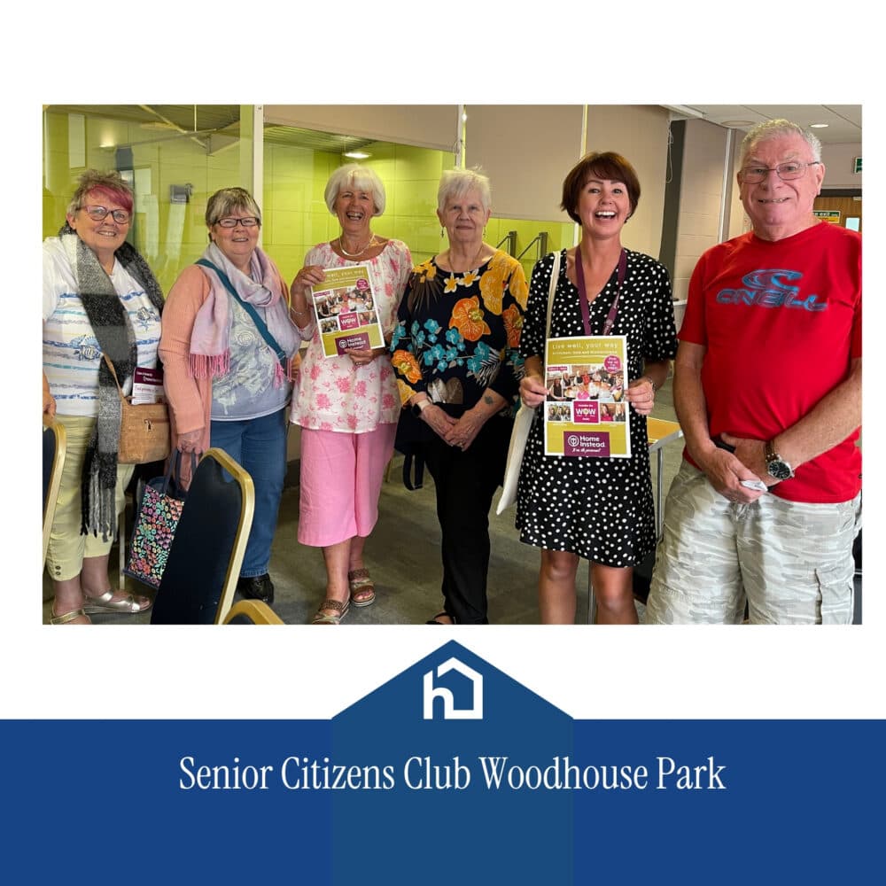 Six smiling pensioners stand together indoors, some holding newsletters, at the Senior Citizens’ Club Woodhouse Park. - Home Instead