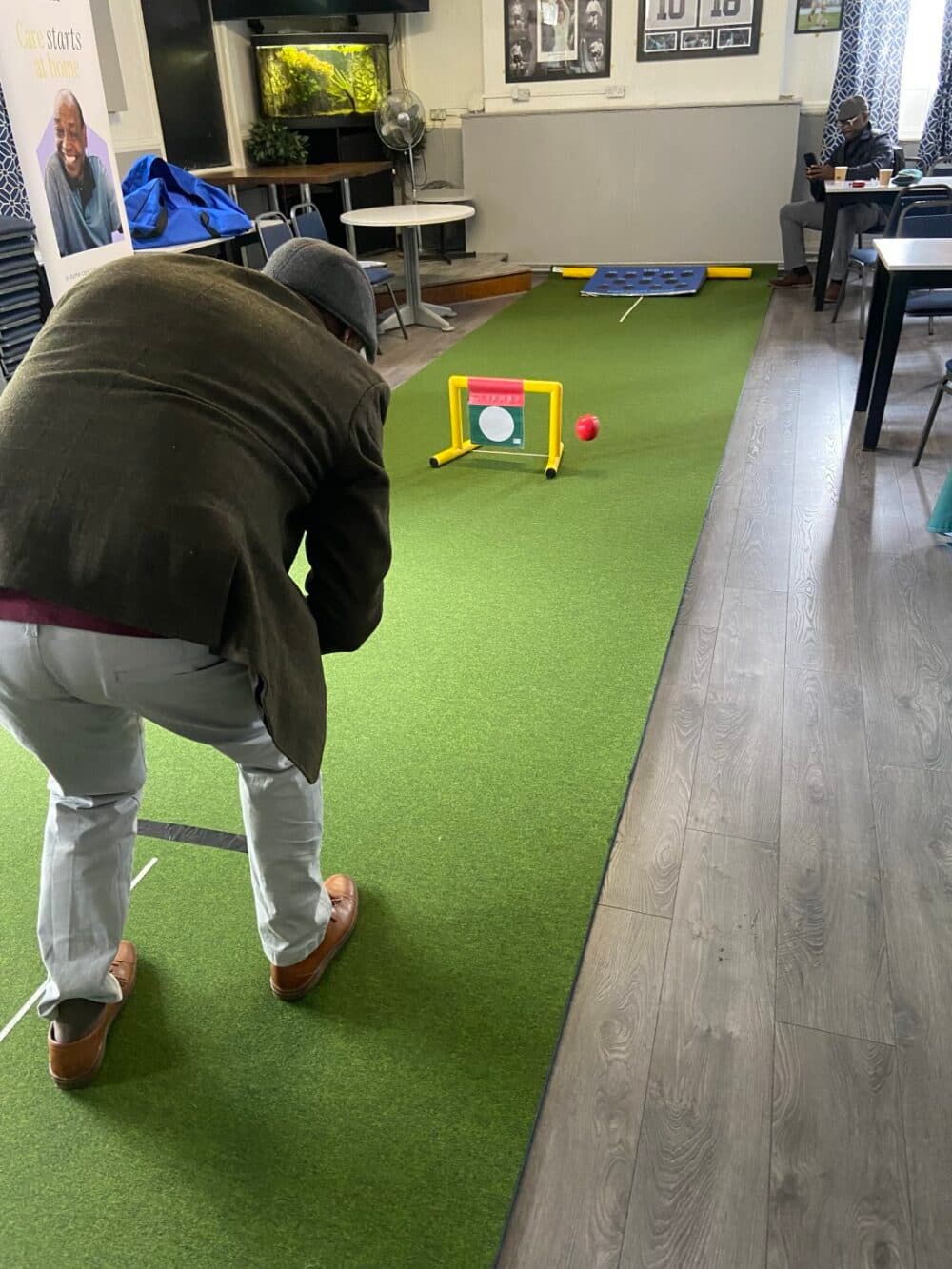 A person plays indoor mini-golf on a green mat in a room with tables, chairs, and wall photos. - Home Instead