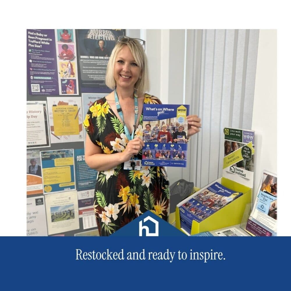 Smiling woman in a floral dress holds a community events magazine in front of an information display. - Home Instead