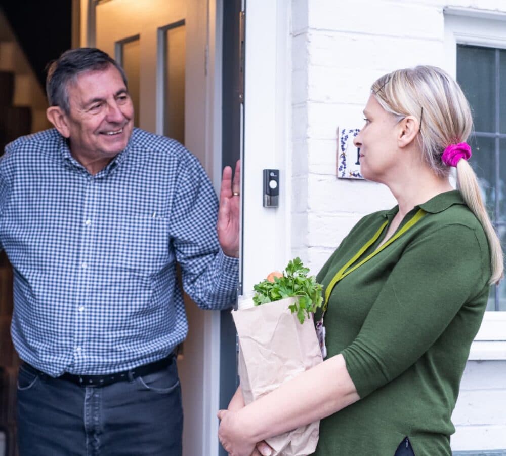 Smiling man greets a woman holding a paper bag of shopping at his front door. - Home Instead
