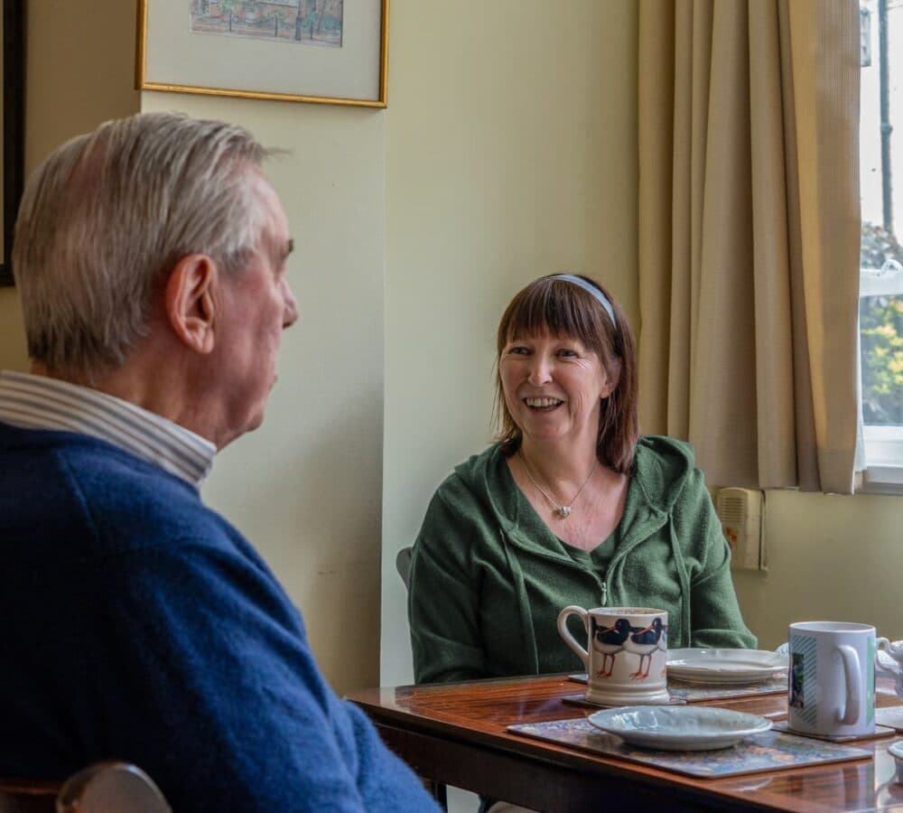 An older male adult with grey hair having tea with his younger female carer with short hair and wearing green inside the restaurant