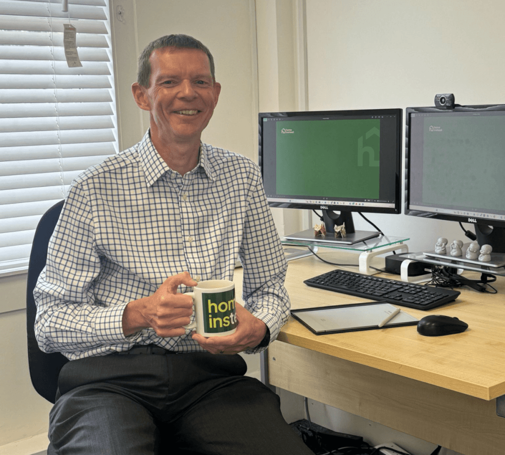 Smiling person in an office holding a mug, sitting at a desk with two monitors and a keyboard. - Home Instead