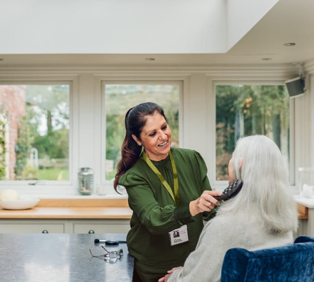 An older female adult with long white hair sitting on a chair while her hair is being combed by her younger female carer happy and smiling inside the house