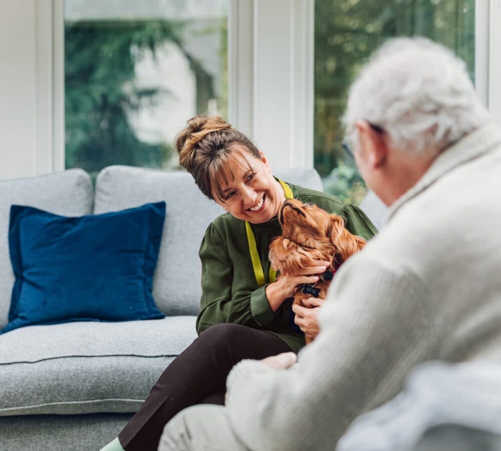 A woman holding a brown dog while sitting on a couch and chatting with an older male adult inside the house