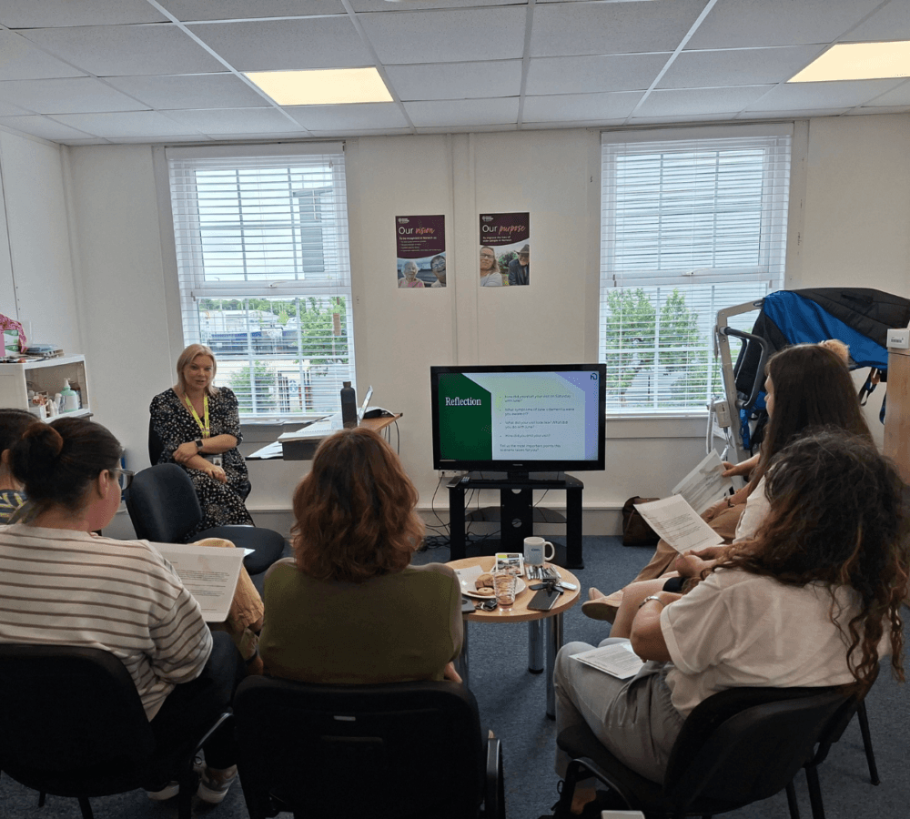 Six women sit in an office, watching a presentation on a screen with notes and refreshments on the table. - Home Instead