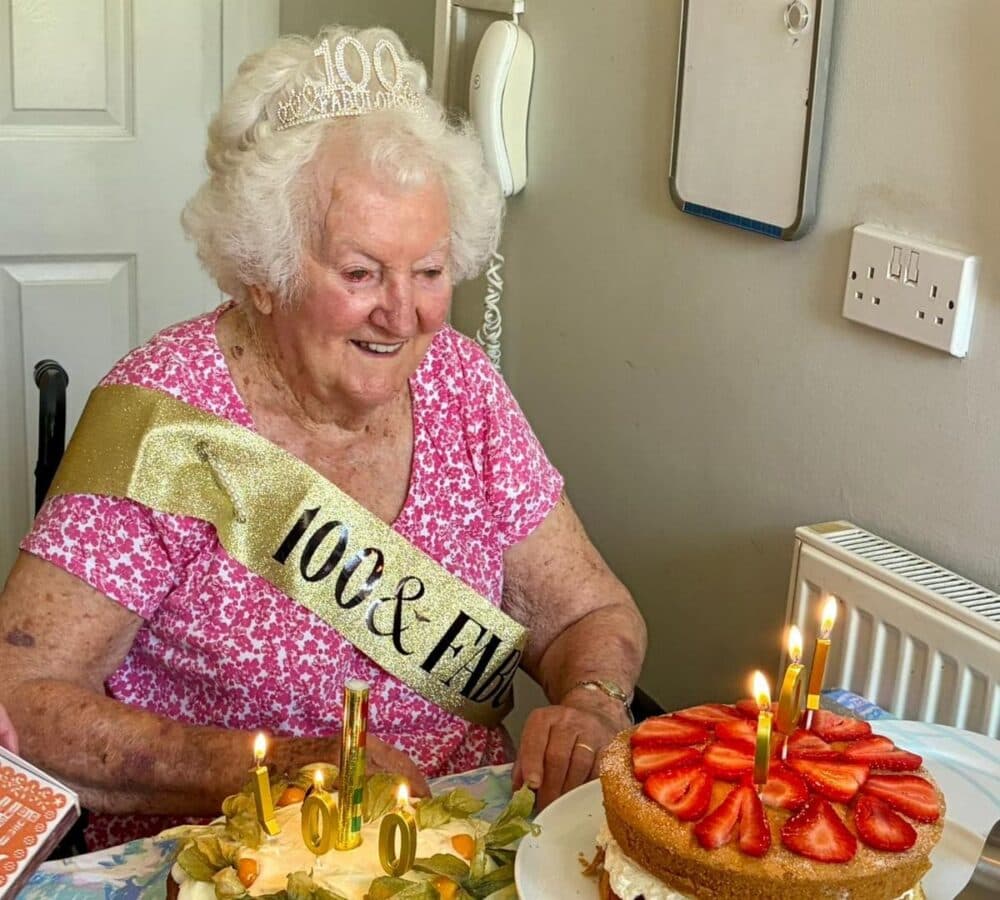 A woman wearing pink and a sash and a crown and with cakes on the table while celebrating her 100th birthday