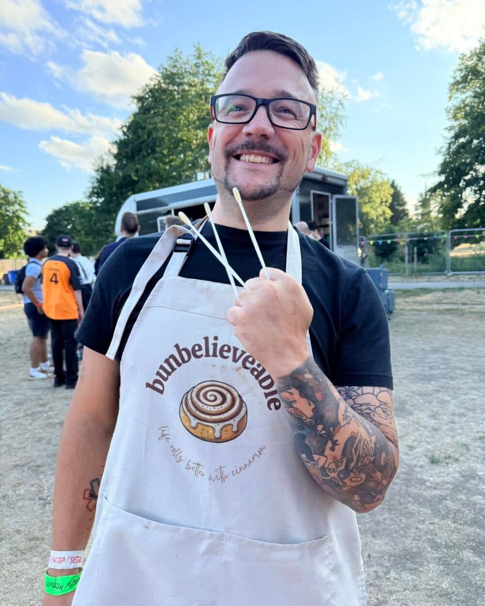 Smiling person in a "bunbelievable" apron holds baking sticks outdoors at a festival or fete. - Home Instead