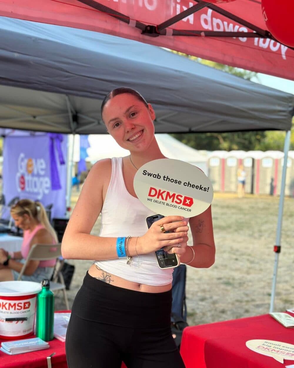 Smiling woman under a marquee holds a DKMS sign promoting cheek swabs for blood cancer awareness. - Home Instead