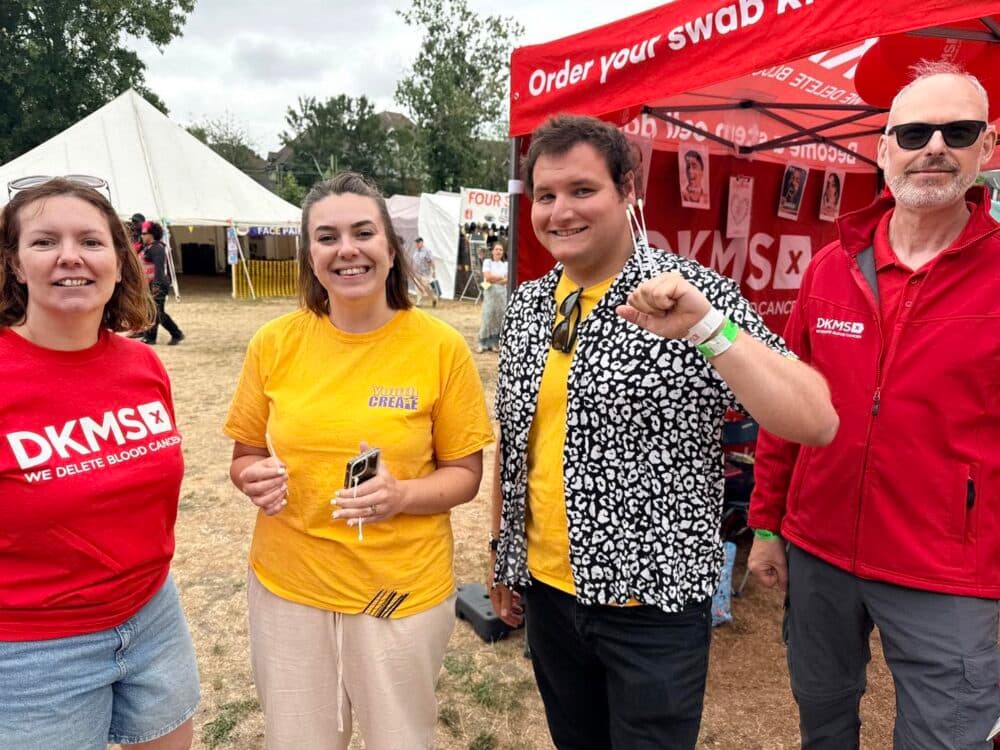 Four people smiling at an outdoor event near DKMS and festival marquees, wearing casual and branded clothes. - Home Instead