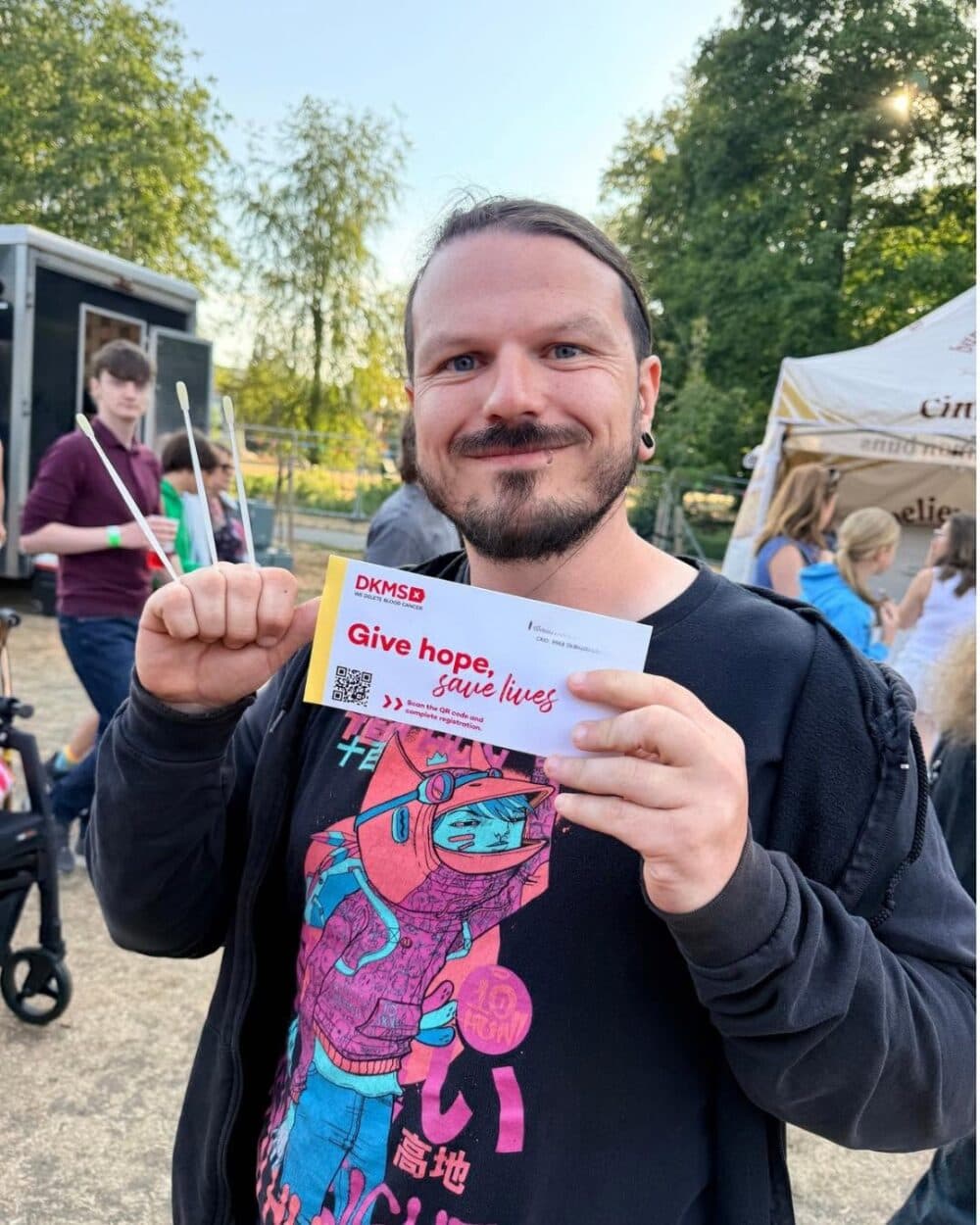 Man holding a "Give hope, save lives" DKMSB card at an outdoor event, smiling at the camera. - Home Instead
