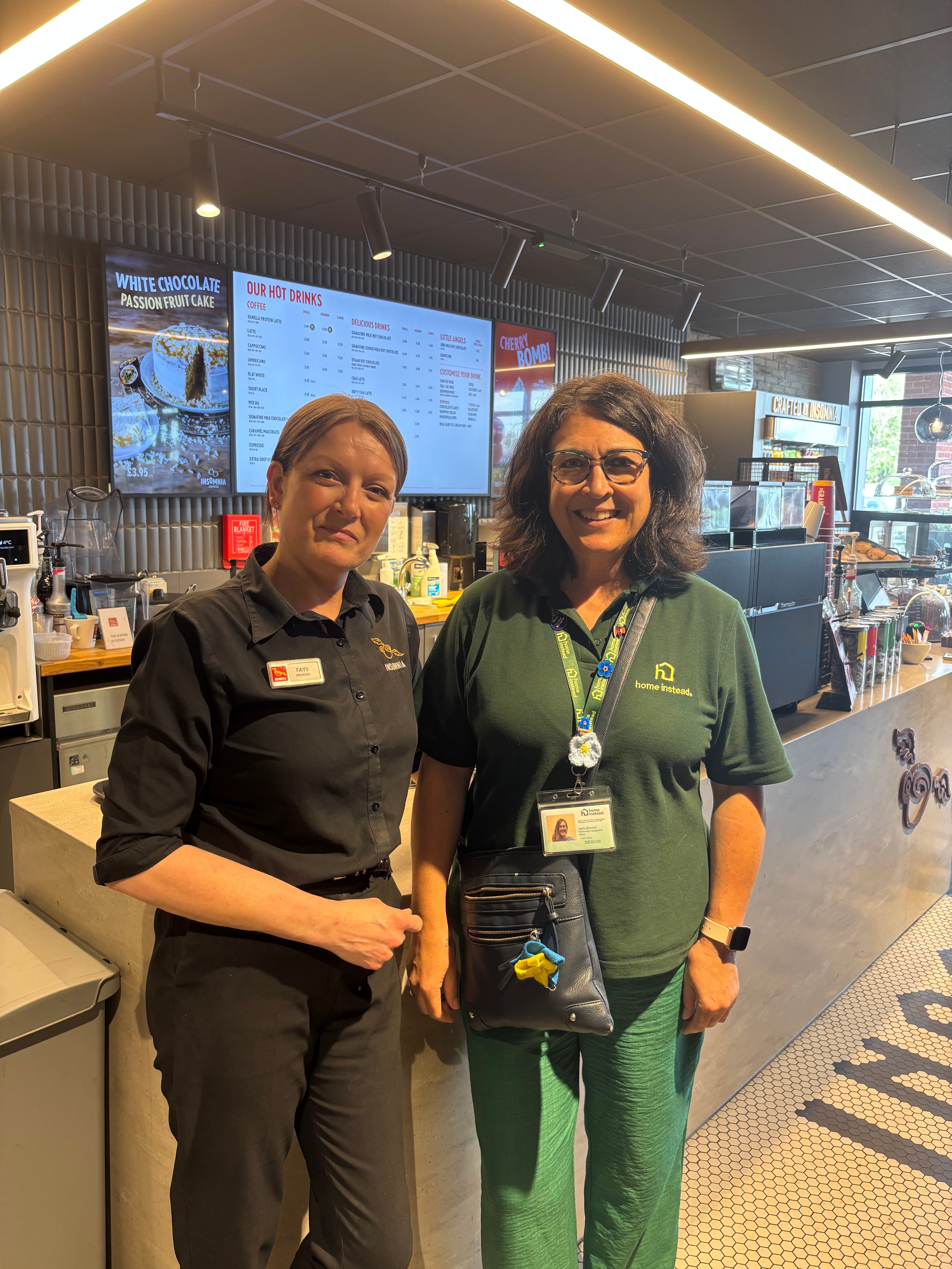 Two women smiling and standing together inside a modern café near the counter, with menus on the wall behind them. - Home Instead