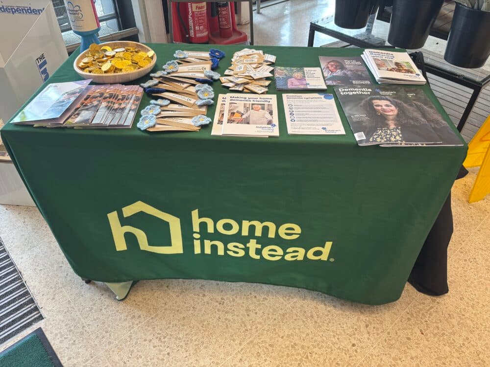 A green "Home Instead" table with leaflets, brochures, pens, badges, and sweets on display. - Home Instead