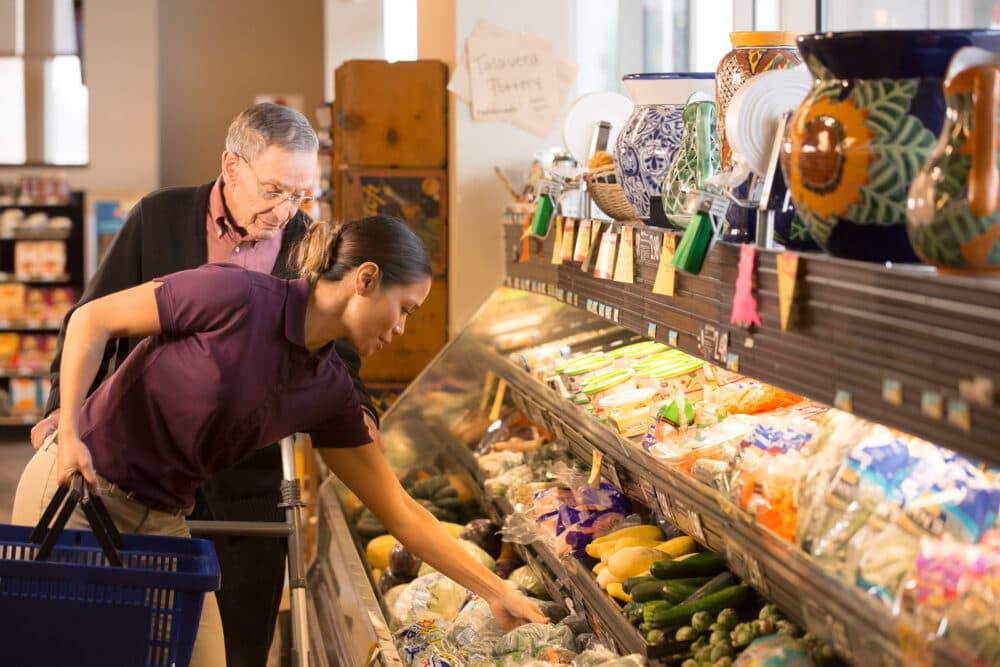 Young female carer shopping with an elderly man, selecting vegetables at the fruit and vegetable aisle in a supermarket