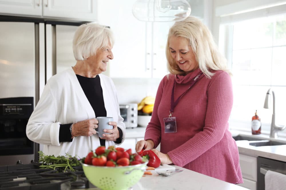 Female carer in her client's kitchen, chatting to elderly female client who is holding a mug, whilst cutting vegetables to prepare a meal
