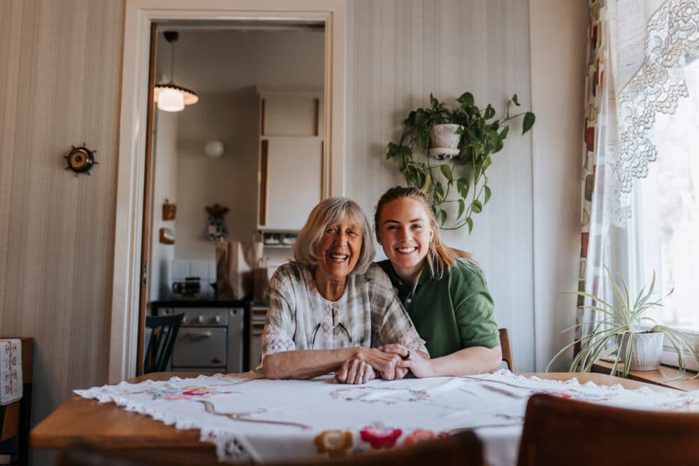 Two women smiling and sitting together at a kitchen table with plants and lace curtains nearby. - Home Instead