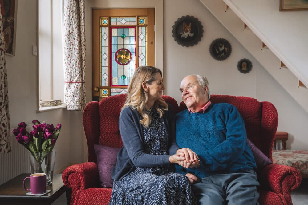 Smiling woman and elderly man sit on a red settee holding hands in a cosy, warmly lit living room. - Home Instead