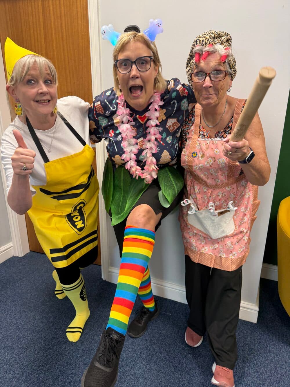 Three women in fancy dress pose together, smiling; one wears a crayon outfit, one is in rainbows, one in an apron. - Home Instead