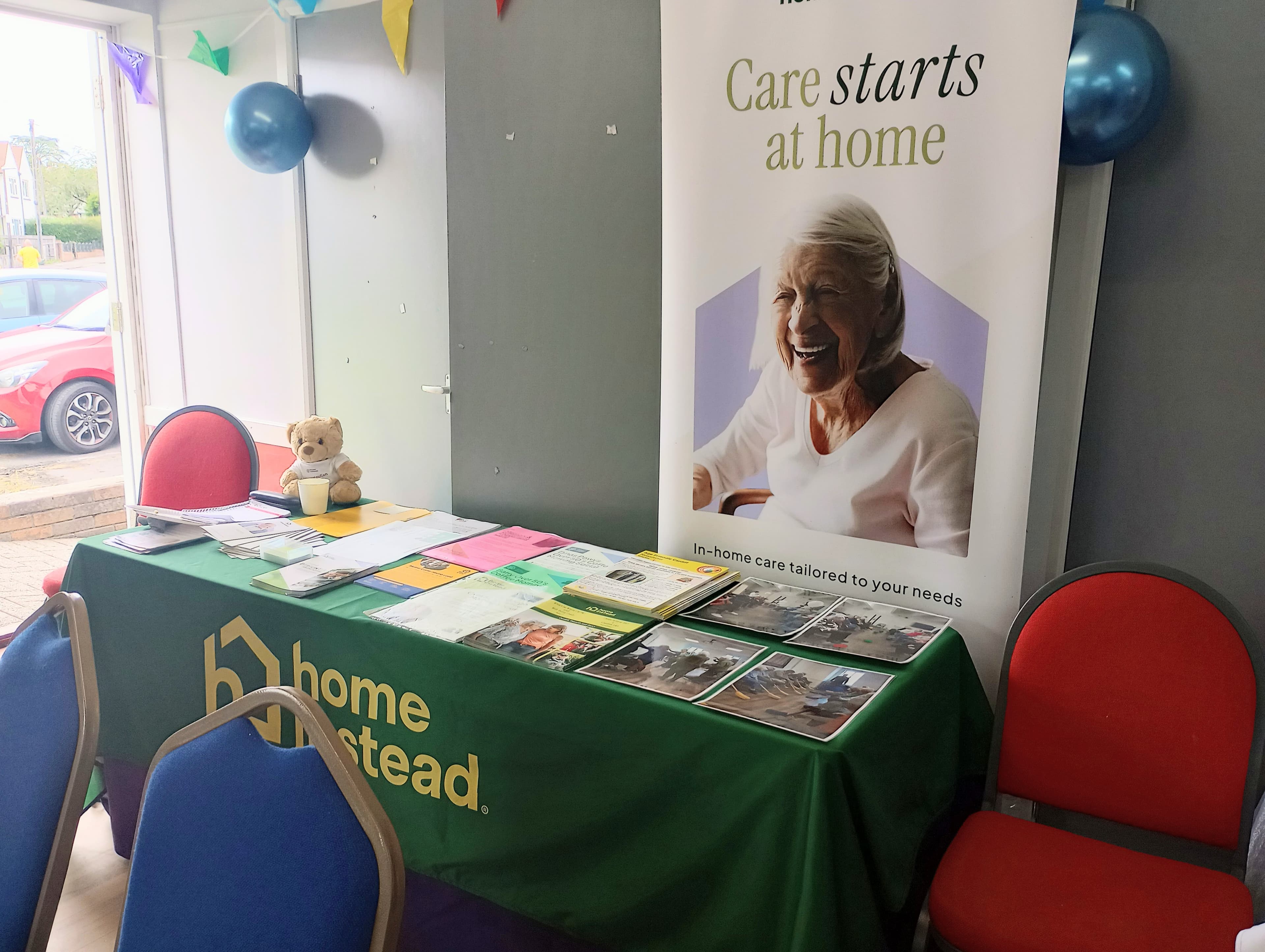 Display table with brochures, a teddy bear, and a banner about home care, decorated with balloons. - Home Instead