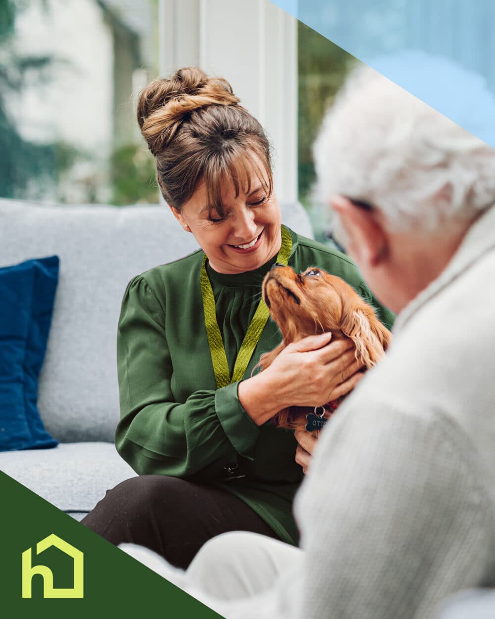 A smiling woman with a lanyard holds a brown dog, sitting with an older man on a sofa. - Home Instead