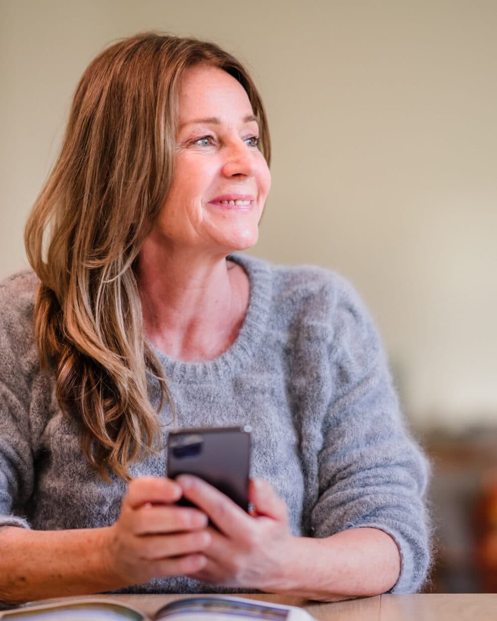 Smiling woman with long hair holding a mobile phone, seated at a table with an open book in front of her. - Home Instead