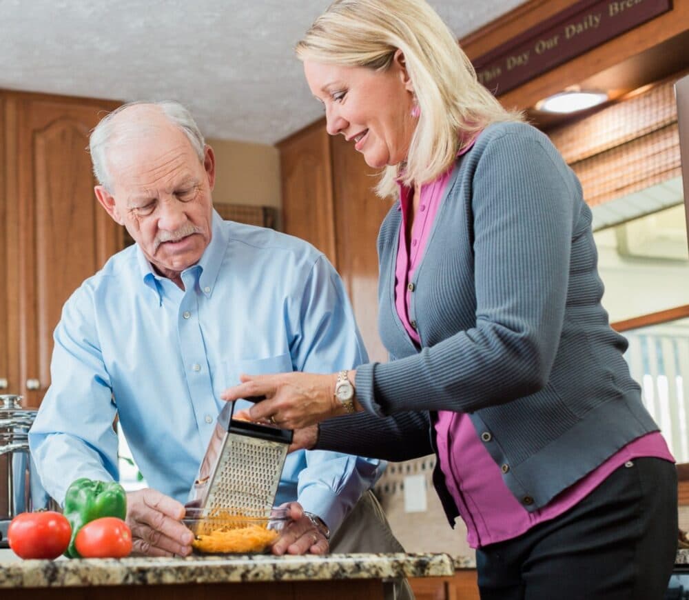 Younger woman and elderly man in the kitchen preparing a meal together. The woman is grating cheese into a glass bowl whilst the man is holding the bowl.